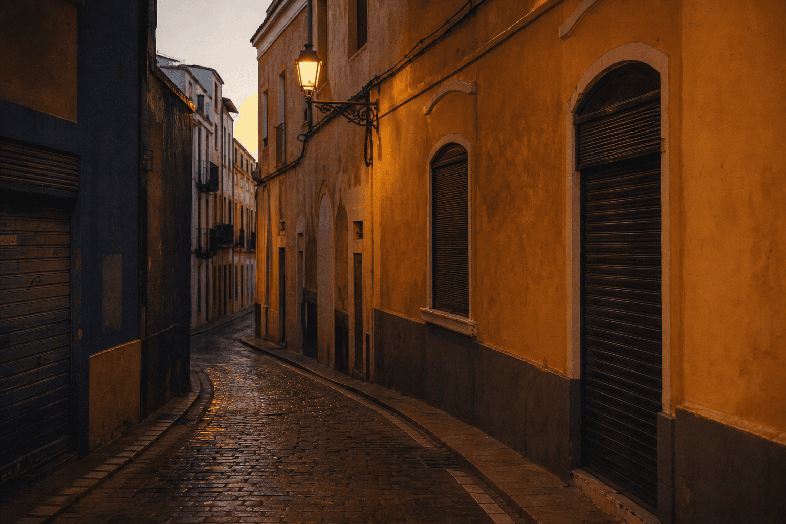 An empty street in a warm southern city at dusk, one amber lamp lit